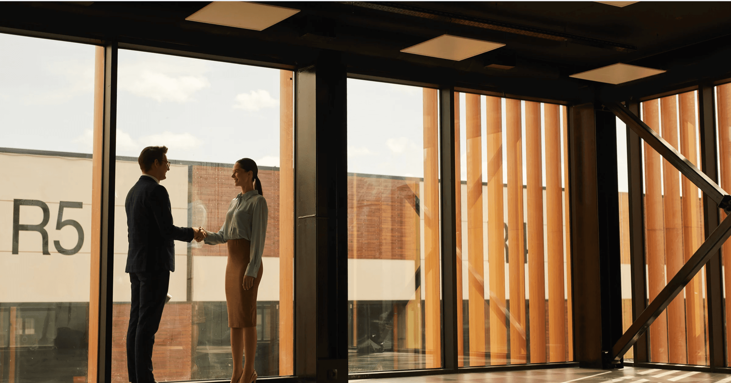 two professionals shaking hands in a glass-walled office overlooking a modern development